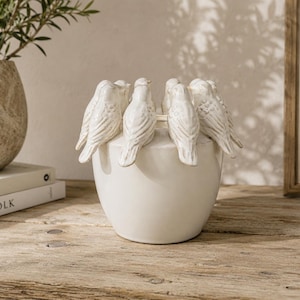 May include: A white ceramic bowl featuring a decorative rim adorned with several sculpted bird figures. The birds are arranged in a circle around the top edge of the bowl, each facing outward with detailed feather textures and a smooth finish.