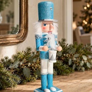 May include: A nutcracker figurine featuring a bright blue glitter finish, a tall hat, and white hair. The figure wears a blue jacket with silver accents and white trousers, holding two silver drumsticks over a small white drum with silver trim.