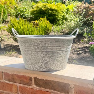 May include: A galvanized metal basin with a mottled silver-gray finish, featuring a wide circular opening, a tapered body with horizontal ridges, and two small, curved handles attached to the upper rim on opposite sides of the container.