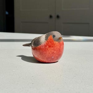 May include: A small, rounded ceramic bird figurine featuring a bright red breast and a grey head and back. The bird has a small, dark eye and a tiny, pointed orange beak. The surface of the figurine has a smooth, glossy finish throughout.