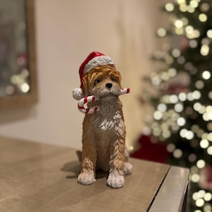 May include: A brown and white dog ornament wearing a red and white Santa hat and holding a candy cane. The dog is sat on a wooden surface. A Christmas tree is visible in the background.