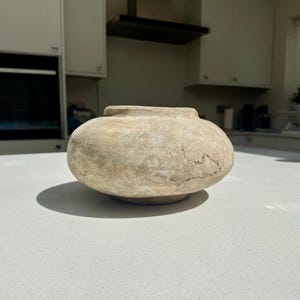 May include: A low, wide, round ceramic vessel with a weathered, light beige and grey textured surface. The bowl features a short, narrow neck with a flat rim and a visible crackled finish across its exterior, resting on a small, circular base.