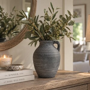 May include: A dark grey, textured ceramic vase with a single side handle, containing a cluster of green olive branches with small berries. The vase has a rounded body and a wide neck, showcasing a weathered, matte finish with visible horizontal ridges.