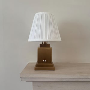 Pode incluir: Um candeeiro de mesa com uma base quadrada de latão escovado e um pequeno botão circular na parte frontal. O candeeiro é rematado por um abajur de tecido branco plissado de forma cónica que se alarga na base, com um aspeto estruturado.