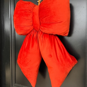 May include: A large, plush, red velvet bow, possibly for festive decoration, hangs against a black door. The bow is richly textured, with a prominent knot and flowing tails. The vibrant colour and substantial size make it a striking decorative piece.
