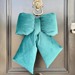 May include: A large, teal velvet bow hangs on a black door. The bow is plush and has a classic bow shape. A gold lion-head door knocker is above the bow.