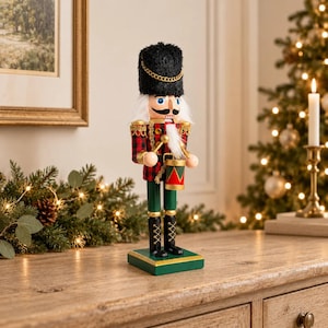 Puede incluir: Una figura de cascanueces de madera con un sombrero negro alto y difuso con una cadena dorada, una chaqueta de cuadros rojos y negros y pantalones verdes. La figura sostiene dos baquetas y un pequeño tambor rojo y verde sobre una base verde.
