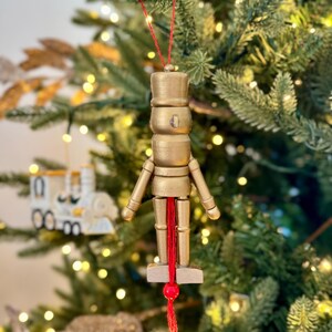以下が含まれることがあります： 金色の木製くるみ割り人形のクリスマスオーナメント。赤い紐が付いており、吊り下げられます。帽子と腕を持つ兵士の形をしています。背景はライトの付いたクリスマスツリーです。