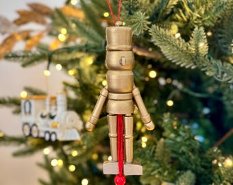 May include: A gold-painted wooden nutcracker Christmas ornament with a red string for hanging. The ornament is in the shape of a soldier with a hat and arms. The background is a Christmas tree with lights.