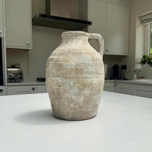 May include: A weathered, light beige ceramic jug with a textured, matte finish. The vessel features a rounded body, a short neck, and a single side handle. The surface displays subtle horizontal ridges and a distressed, aged appearance throughout.