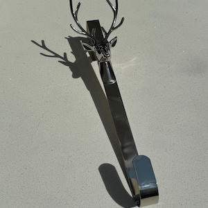 Könnte beinhalten: Ein silberfarbener Strumpfhalter aus Metall mit einem dreidimensionalen Hirschkopf und detailliertem Geweih an der Oberseite. Das Design umfasst einen langen, flachen vertikalen Arm, der nach unten verläuft und in einem gebogenen Haken endet.