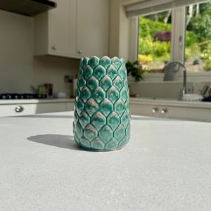 May include: A cylindrical ceramic vase featuring a repeating scale-like pattern across its exterior. The surface is finished in a glossy teal glaze with subtle brown highlights on the edges of each individual scale, creating a textured appearance.