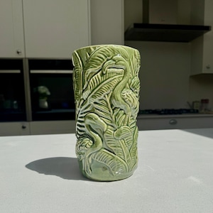 Peut inclure: Un mug tiki en céramique verte avec un motif en relief détaillé de flamants roses et de feuillage tropical. Le mug a une forme cylindrique et une finition brillante. Le motif enveloppe tout le mug, créant une surface texturée.