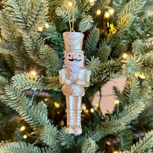 Puede incluir: Adorno navideño de cascanueces dorado. El adorno tiene un diseño detallado, que incluye un sombrero, un bigote y un uniforme. Cuelga de un hilo dorado sobre un fondo de un árbol de Navidad verde con pequeñas luces.