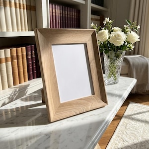 May include: A rectangular picture frame made of light-coloured wood with a visible grain pattern. The frame features a wide, bevelled border surrounding a blank white rectangular insert, designed to stand upright on a flat surface.
