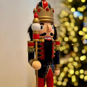 May include: A wooden nutcracker figurine, with a red, black, and gold colour scheme. The figurine wears a crown and holds a staff. The background is blurred, with a Christmas tree visible. The figurine is a festive decoration.