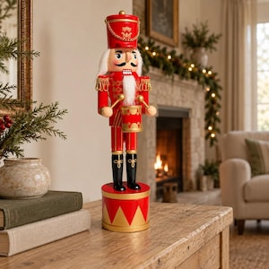 May include: A wooden nutcracker figure dressed in a bright red uniform with gold trim, a tall hat, and black boots. The figure holds two drumsticks and stands on a red cylindrical base with gold triangular accents, playing a small red drum.