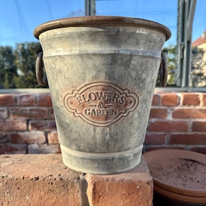 Puede incluir: Un cubo de metal galvanizado con un acabado envejecido, con el texto en relieve "FLOWERS & GARDEN". El cubo tiene dos asas pequeñas y un borde enrollado. Está sobre una superficie de ladrillo rojo.