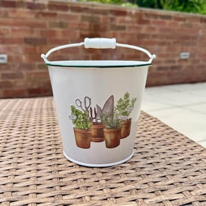 Könnte beinhalten: Ein weißer Garten-Metalleimer mit grünem Rand und weißem Griff. Der Eimer zeigt Illustrationen von Gartengeräten und Topfkräutern, darunter Basilikum und Rosmarin. Der Eimer steht auf einer gewebten braunen Oberfläche.