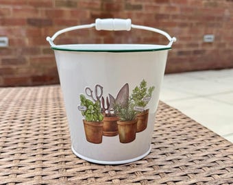 Pode incluir: Um balde de jardim de metal branco com uma borda verde e uma alça branca. O balde apresenta ilustrações de ferramentas de jardinagem e ervas em vasos, incluindo manjericão e alecrim. O balde está em uma superfície marrom tecida.