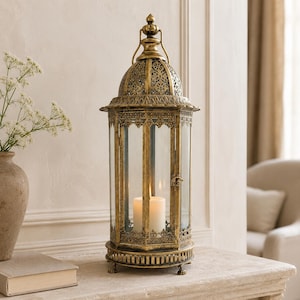 Può includere: Una lanterna esagonale alta con finitura oro anticato, caratterizzata da intricati intagli in metallo sulla cupola e sulla base. La lanterna presenta pannelli in vetro trasparente e una piccola chiusura in metallo laterale con una candela.