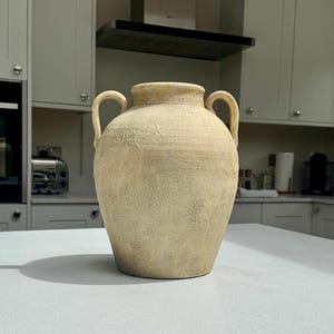May include: A beige ceramic vase with a textured, weathered finish, featuring a rounded body, a short neck, and two symmetrical handles on either side. The surface has a mottled appearance with subtle variations in tone and a matte texture.