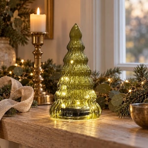 May include: A translucent olive green glass Christmas tree figurine with a ribbed, wavy surface texture. The hollow interior contains a string of warm white LED fairy lights that illuminate the glass structure from within, creating a soft glow.