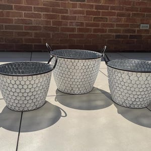 May include: Three round metal buckets with a white honeycomb pattern and black rims. Each bucket features two black metal handles on opposite sides. The buckets have a tapered shape, narrowing toward the base, and are arranged in a triangular formation.
