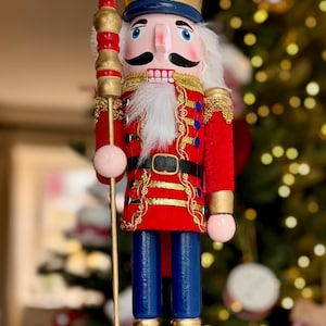 Puede incluir: Figura decorativa de cascanueces de madera con temática navideña. El cascanueces viste una chaqueta roja con ribetes dorados, pantalones azules y botas negras. Tiene una corona dorada, barba blanca y sostiene un bastón dorado con una esfera rosa.