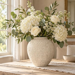 Könnte beinhalten: Eine runde, texturierte, cremeweiße Keramikvase enthält ein großes, üppiges Blumenarrangement. Der Strauß besteht aus weißen Hortensienblüten, cremefarbenen Rosen, kleinen weißen Füllblumen sowie verschiedenen grünen Eukalyptusblättern.