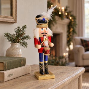 May include: A decorative nutcracker figurine wearing a red jacket with gold trim, blue trousers, and a black hat adorned with a white feather and a blue gem. The figure holds two gold drumsticks and stands on a square gold glittered base.