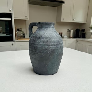 May include: A dark gray, weathered ceramic jug with a single side handle. The surface features a textured, matte finish with subtle horizontal ridges and a distressed, uneven appearance throughout the body, narrowing toward the top opening.