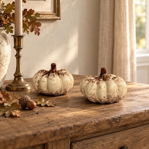 May include: Two decorative ceramic pumpkins with a glossy cream-coloured finish and brown distressed detailing. Each pumpkin features a smooth, rounded shape with vertical ridges and a dark brown stem, sitting on a wooden surface.