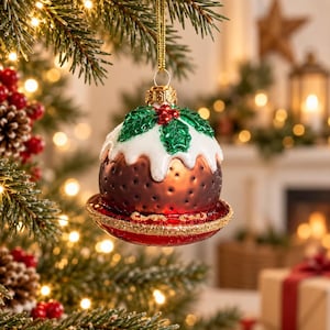 May include: A glass Christmas tree ornament shaped like a traditional plum pudding. The ornament features a dark brown base with a white icing top, green holly leaves, and small red berries. It sits on a red plate with a gold glittered rim.