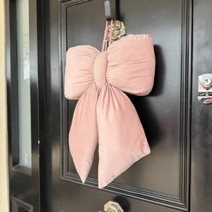 May include: A large, plush, pink velvet bow hangs on a black door. The bow is made of soft, gathered fabric and is suspended by a pink cord from a metal hook. The door has a brass doorknob.