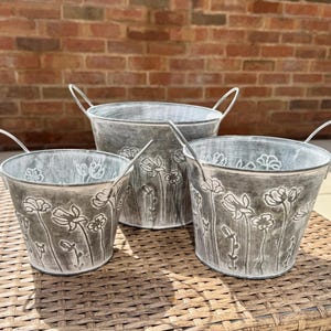 May include: Three galvanised metal flower pots with handles. The pots are grey with a distressed finish and a floral design. The pots are different sizes and are arranged on a woven surface.