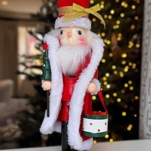 May include: A festive Christmas nutcracker in the shape of Santa Claus. The wooden figure wears a red coat with white trim, a red hat, and holds a small green Christmas tree. A gold bow adorns the hat.