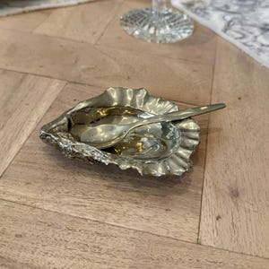 May include: A silver oyster shell dish with a small spoon inside. The dish is on a wooden table.