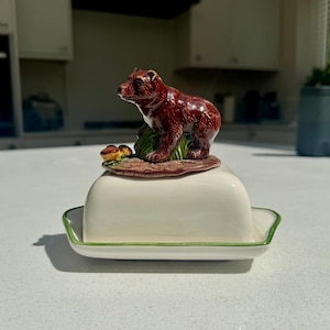 May include: A ceramic butter dish featuring a glossy off-white rectangular base with a thin green rim. The lid is topped with a sculpted, hand-painted brown bear standing on a patch of green grass and small brown mushrooms, creating a decorative handle.