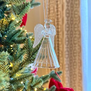 Puede incluir: Adorno navideño de ángel de cristal transparente con alas plateadas brillantes y un halo, colgado de un hilo plateado. El ángel está suspendido frente a un árbol de Navidad decorado con lazos rojos y luces.