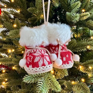 May include: A pair of red and white Christmas boot ornaments with white faux fur trim and small white pom-poms. The boots are decorated with a snowflake pattern and are hanging from a Christmas tree.