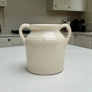 May include: A cream-coloured ceramic vessel featuring a wide mouth, two curved side handles, and a subtle embossed dotted band encircling the upper portion of the body. The surface has a glossy, slightly speckled finish with a smooth texture.