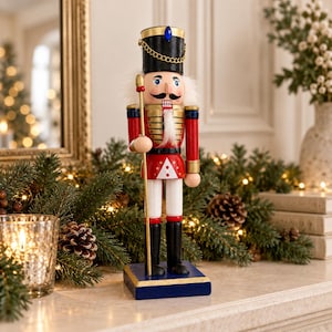 Könnte beinhalten: Eine hölzerne Nussknacker-Figur auf einem blauen Sockel, die einen schwarzen Hut mit blauem Edelstein und goldener Kette, eine rote Jacke mit Goldbesatz, weiße Hosen und schwarze Stiefel trägt, einen goldenen Stab hält und einen weißen Bart hat.
