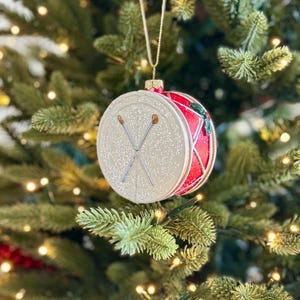 Può includere: Un ornamento per l'albero di Natale a forma di tamburo. L'ornamento ha una faccia bianca e glitterata con bacchette incrociate e un lato rosso e glitterato. È appeso a un filo d'oro su uno sfondo di albero di Natale verde.