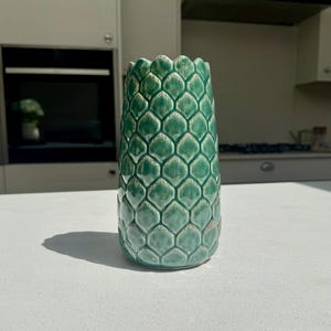 May include: A cylindrical ceramic vase featuring a textured, repeating scale pattern in shades of emerald and sage green. The surface has a glossy finish with subtle white highlights along the edges of each individual scale-like protrusion.