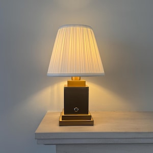 May include: A table lamp with a gold-toned square base and a white pleated fabric lampshade. The base features a small circular power button on the front. The lamp emits a warm glow, illuminating the shade and casting light onto the surface below.
