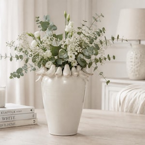 Puede incluir: Un jarrón de cerámica de color blanco roto brillante con un borde decorativo adornado con una serie de pequeñas figuras de pájaros esculpidas. El jarrón está lleno de un exuberante arreglo de ramas de eucalipto verde, flores de lisianthus blancas y paniculata.