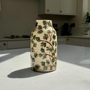 May include: A ceramic vase with a cream-coloured base featuring a crackle glaze finish. The surface is decorated with a hand-painted pattern of brown branches and small green leaves that wrap around the cylindrical body of the vessel.