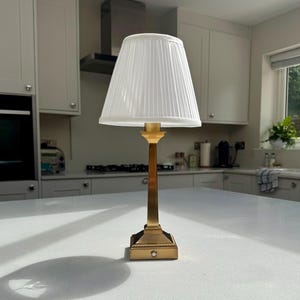May include: A table lamp featuring a tall, slender, brushed gold-tone metal base with a square, beaded-edge pedestal and a white, pleated, tapered fabric lampshade. The lamp has a small, circular power button located on the front of the base.