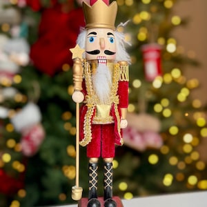 Peut inclure: Figurine décorative en bois de casse-noisette. La figurine porte une veste en velours rouge avec des bordures dorées, une couronne dorée et une barbe blanche. Il tient un bâton doré avec une étoile. Le casse-noisette repose sur une base rouge.